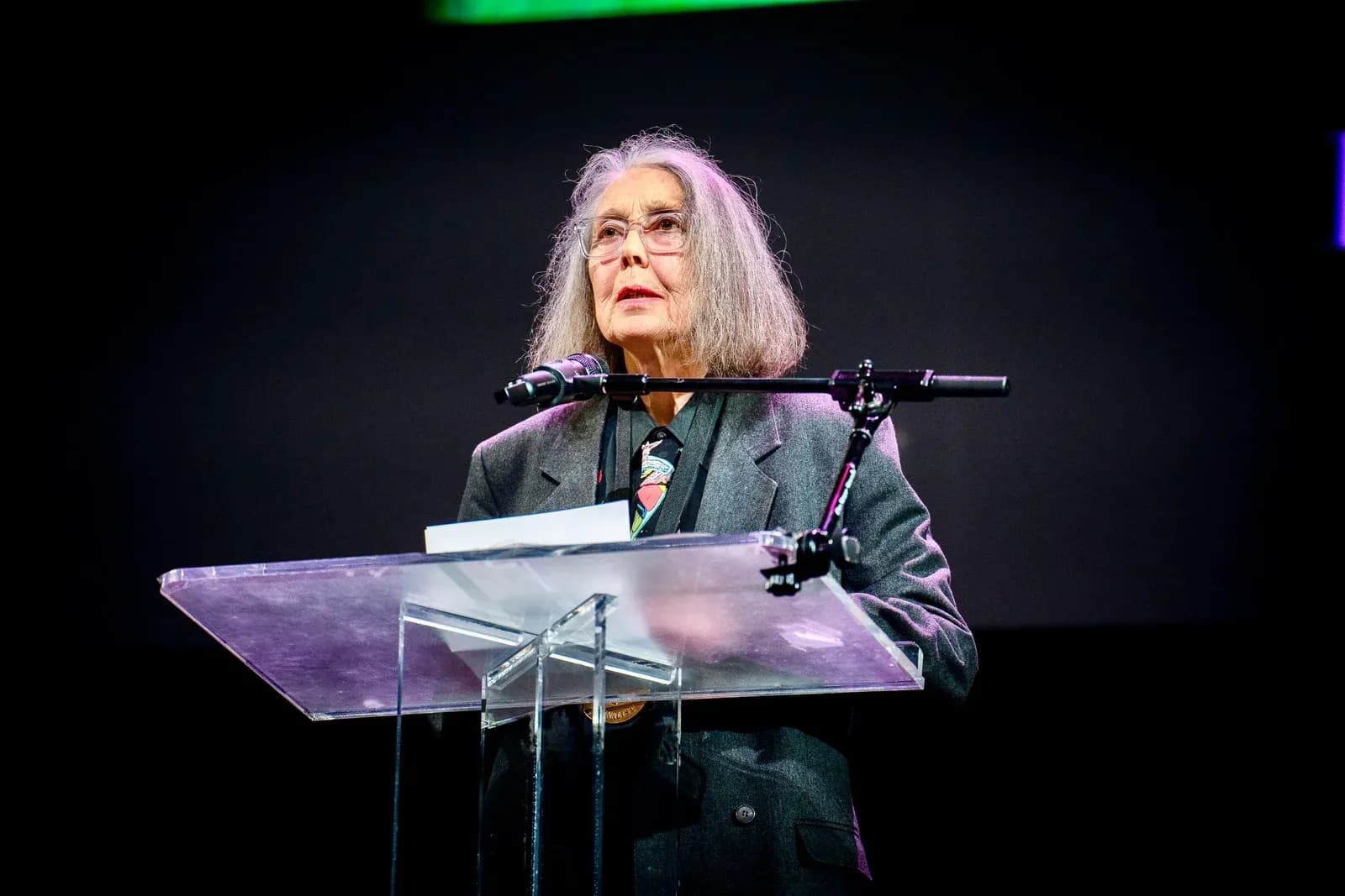 Anne Carson, in glasses and a grey jacket, speaks at a clear acrylic lectern at the 2024 National Book Awards finalist reading in New York.
