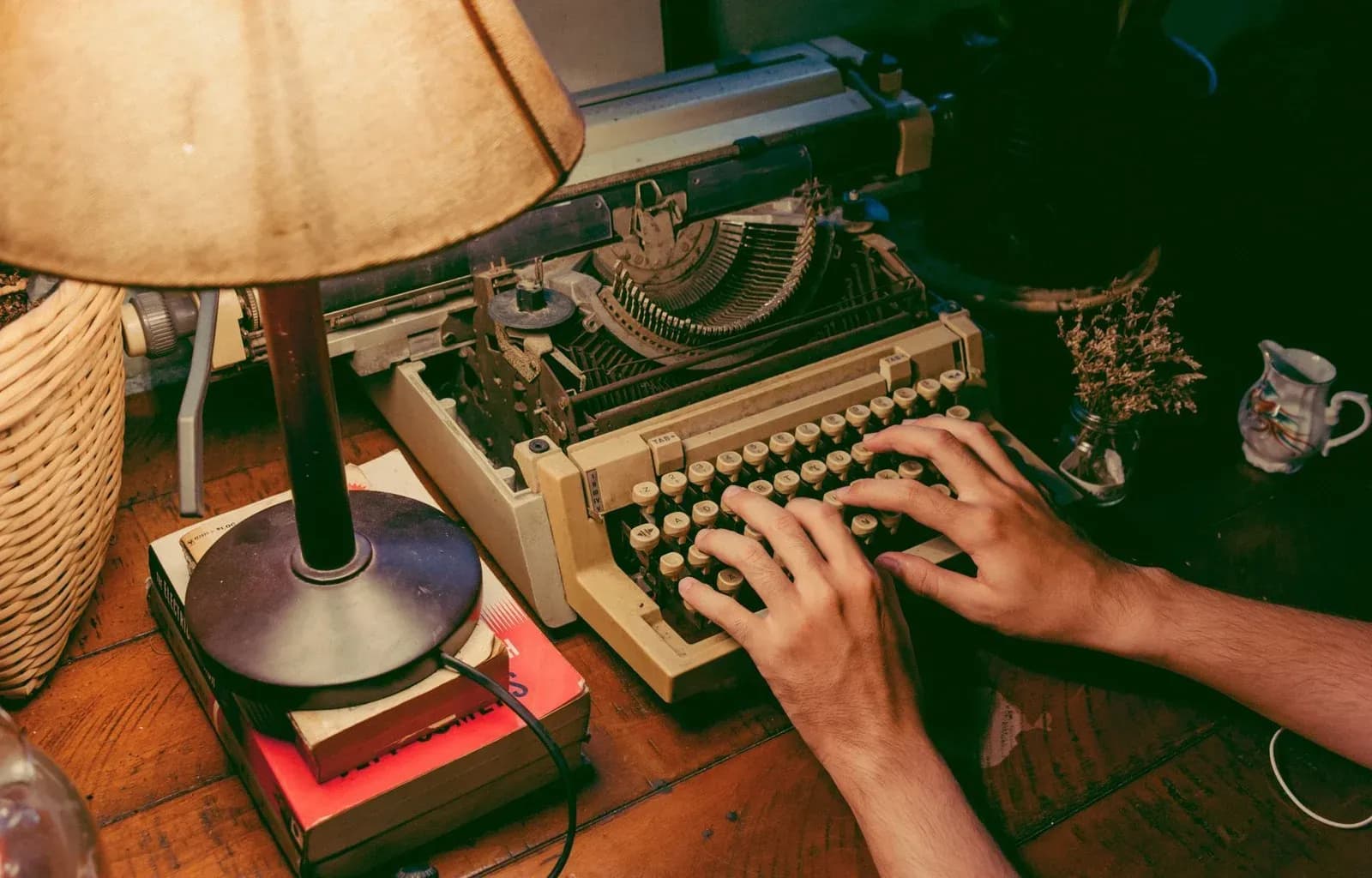 A person types on a vintage typewriter at night under the light of a desk lamp.