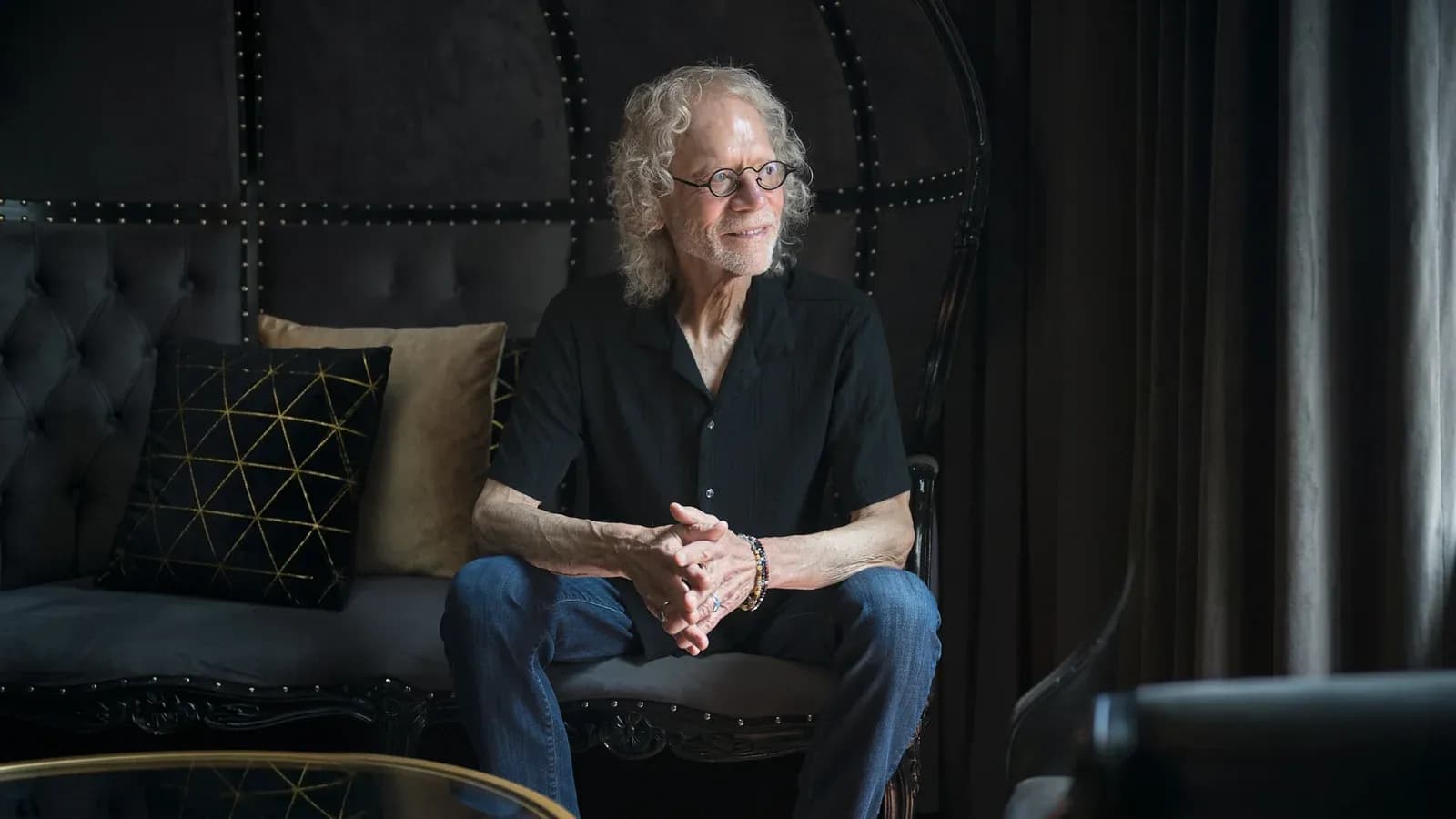 Tom LeNoble seated on a sofa in a dark, studded interior, hands clasped